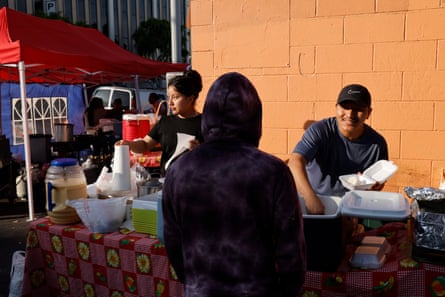 two people stand at table outside and give food to a person