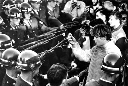A man places a flower into a gun barrel as he’s surrounded by law enforcement officers