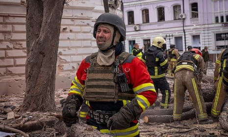A rescue worker in helmet, body armour and high-visibility clothing against a background of other emergency workers amid debris