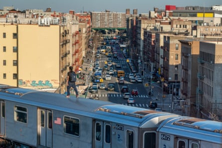 A person stands atop a moving subway train over a busy NYC street