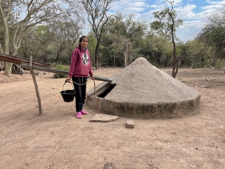 A woman stands by a covered well in a dry, sandy patch with trees in the background