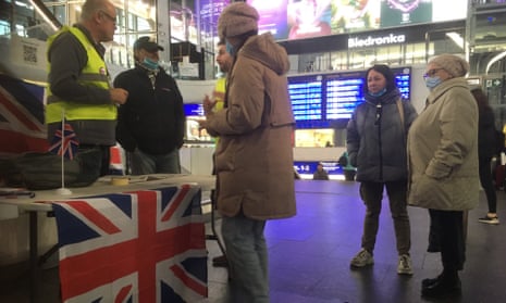 Two men stand at a stall with a union jack draped over it in a train station talking to people