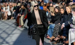 A model walks down the catwalk in a black outfit, which includes an unbuttoned jacket exposing bare flesh