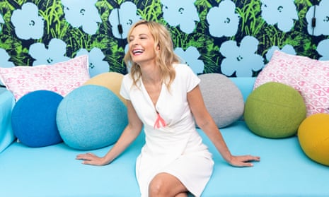 Wednesday Martin in a white dress and wearing a clitoris shaped necklace, sitting on a pale blue sofa with round coloured cushions