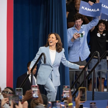 harris climbs stairs, smiling, in front of people holding up ‘kamala’ signs