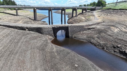 Dry reservoir with only a small channel of water in the centre deep below two bridges.
