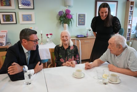 Andy Burnham sits at a table with two elderly residents while a staff member stands nearby in a care home