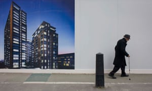 An elderly man walks bent past a regeneration project hoarding image at Elephant & Castle, London.
