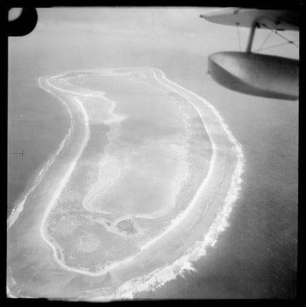 Black-and-white aerial image of an atoll, with part of a bi-plane wing visible