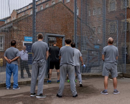 Men wearing grey T-shirts and trousers stand in an exercise yard next to a Victorian building.