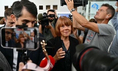 Gisèle Pelicot, smiling slightly, holds up her hand as she is surrounded by cameramen and press microphones