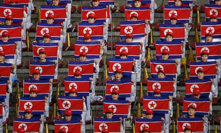 A crowd of people sit in an orderly fashion singing and holding up individual red and blue North Korean flags