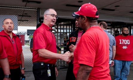 A group of men wear red T-shirts. In the center, a middle-aged white man wearing glasses, Shawn Fain, shakes the hand of a middle-aged Black man who also wears a red baseball cap.