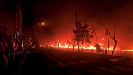 Two people on a motorbike ride past a line of fire ar night.