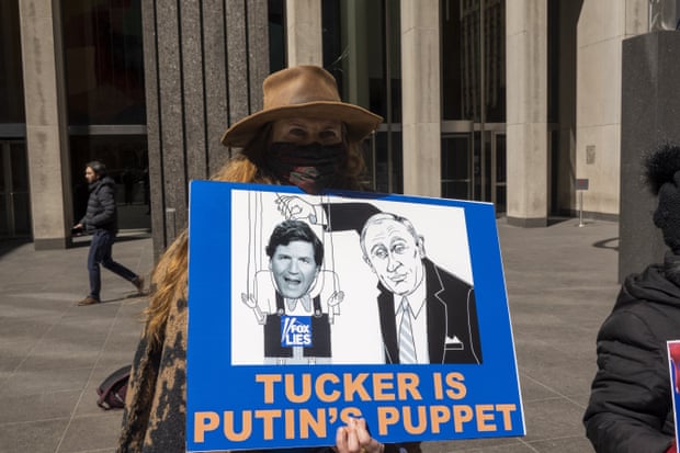 A woman in a hat and protective mask holds a sign with a cartoon depicting Vladimir Putin pulling the strings on a Tucker Carlson puppet. The words below read 'Tucker is Putin's puppet'.