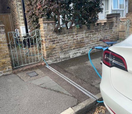 An electric car parked in the road outside a house while charging, with an EV cable channel in the pavement beyond the garden gate and brick wall