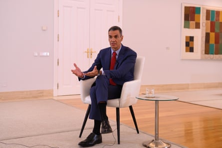 Sánchez sat in a white armchair next to a glass coffee table in a large room with white walls and a colourful print on the wall in the background