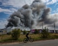 Person rides a bicycle in foreground with Wildberries lorries behind and a big plume of smoke rising into the air