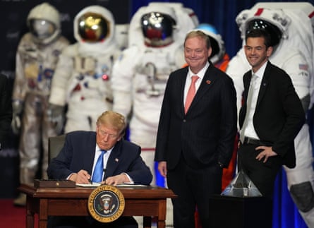 a man signing a document at a desk while two others stand nearby and spacesuits are pictured in the back