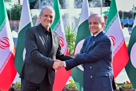 Masoud Pezeshkian, dressed in a black suit, shakes hands with Shehbaz Sharif. Both men are smiling and standing before Iranian and Pakistani flags.