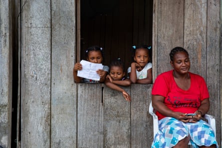 Berta Lozano, mother of nine, in front of her house, with three young girls looking out of a window.