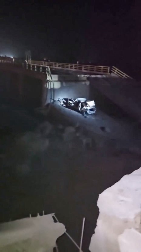 A torch light shines on a burnt our vehicle on a damaged bridge.