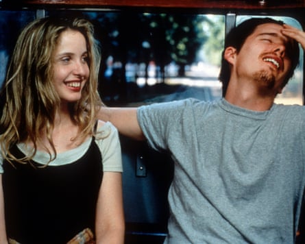 A young woman with uncombed long hair in a T-shirt and vest-dress grins as a smiling young man in a T-shirt, his right arm around her shoulder, throws his head back with his hand on his forehead