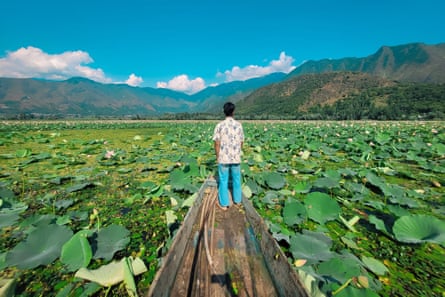 A Kashmiri boy stands in a boat sailing through lotus flowers on Wular Lake, with mountains in the distance.