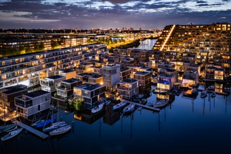 Aerial view at dusk of the floating housing district. Many of the houses are lit up from within and some have small boats moored outside
