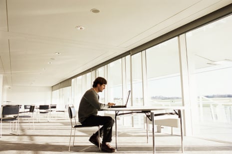 A man sits at a table using a laptop beside a long row of glass windows.