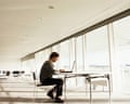 A man sits at a table using a laptop beside a long row of glass windows