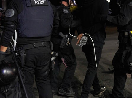Police officers arrest a person wearing all black. Their hands are constricted by white plastic zip ties. 