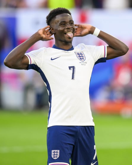 Bukayo Saka stands during a football game with his hands to his ears and his tongue sticking out.