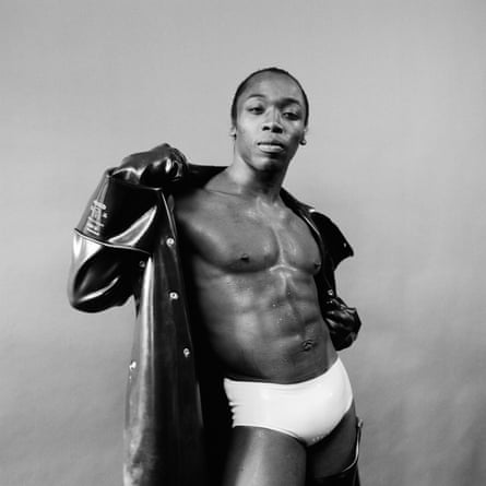 Photograph of a man wearing only white pants and a rubber jacket, which he is holding open to expose his chest and abs