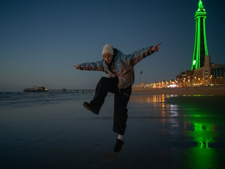 A young man dances in semi darkness on Blackpool beach with the tower outlined in green neon lighting in the background to right of frame.
