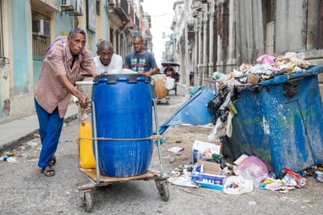 Three men push a large blue drum of water on a trolley through the rubbish-strewn streets of Havana, Cuba.