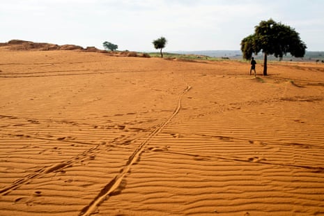 Uma pessoa vista caminhando por uma enorme extensão de areia vermelha com apenas três árvores à vista