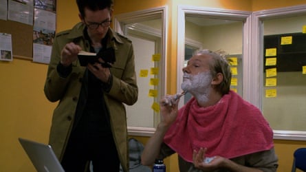 Julian Assange shaving with a man standing next to him with a notebook.