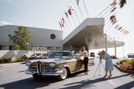 a man poses against a car in front of a hotel with lots of flags as a woman takes a picture in a retro photo