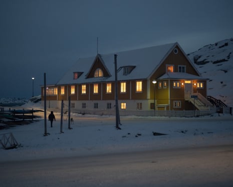 A two-storey building in a snowy landscape with a steeply pitched roof and all its windows aglow with light