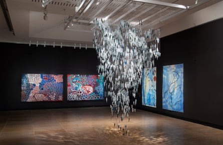 A gallery with black walls, with a series of colourful paintings hung on two of the walls, and suspended from the ceiling in the centre of the room an installation of more than 2000 translucent glass pieces in different shades of blue and grey and shaped like yams, arranged in a formation like rain.