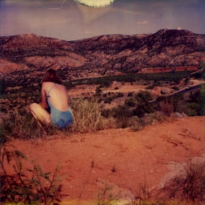A woman in blue swimming costume, her back to us, crouches down in a red-tinged desert, the whole image looking washed-out and nostalgic