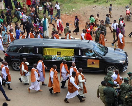 Hearse with coffin draped in flag escorted by guards in orange robes
