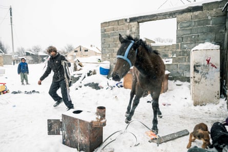 A boy holds a horse by a rope in a snowy landscape. There is a breezeblock wall behind the horse. A younger child stands behind the boy
