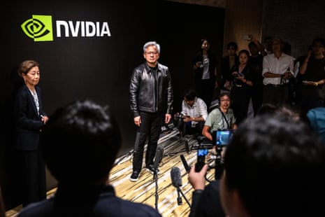 a man in leather jacket and black pants stands in front of a crowd of journalists, a backdrop on the wall behind him saying 'nvidia' with green company logo