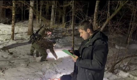 A man writes notes in the foreground, in a clearing of a snowy forest at night. In the background two men inspect the ground near the trees