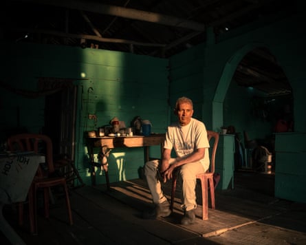 An older man sits on a plastic chair next to a wonky table in a wooden cabin