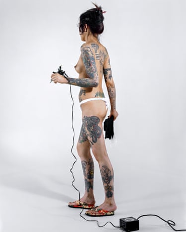 A heavily tattooed, topless woman holds a tattoo machine, with cables trailing to a power unit