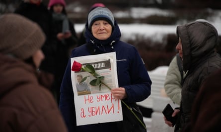An older woman in a knitted hat and winter jacket holding a single rose and holding a piece of paper taped with a newspaper article about Alexei Navalny and words in Russian meaning “Not dead but killed”