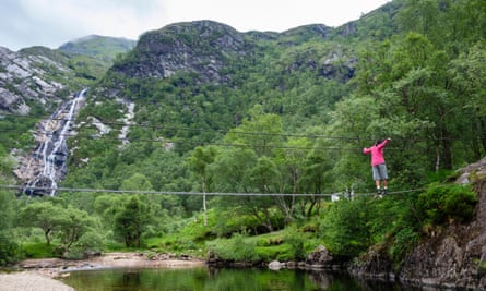 Woman walking across the An Steall wire bridge over the Water of Nevis river with Steall Falls beyond, in Scotland, UK.